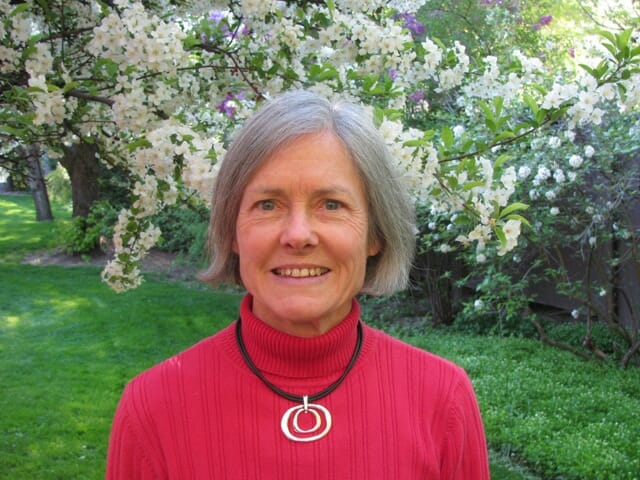 white woman with grey hair in a red sweater about a greenery backdrop