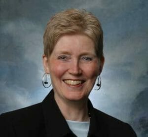 white woman with short light colored hair in a black blazer and grey shirt amid a blue grey backdrop