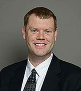 white man with a black suit jacket, white shirt and dark tie amid an olive background