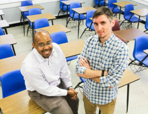 Let to right are a black man in khakis and a white collared shirt and a white man in a checkered shirt and khakis in a classroom