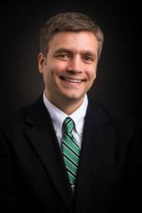 white man with short brown hair, black suit jacket, white shirt, blue and black tie amid a black background