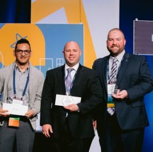left to right three white men receiving their awards. On the left he has shrt dark hair, a grey shirt and dark pants. In the middle he is bald wearing a black suit, white collared shirt and grey tie. To the right he is wearing a dark suit, white collared shirt, and grey tie with short dark hair and a beard.