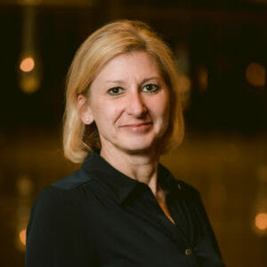 white woman with short blond hair in a black suit with a dark background