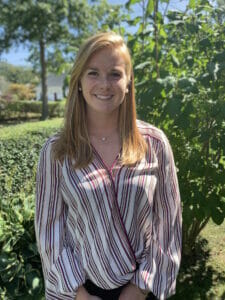 Headshot of a white woman with long blonde hair in a light colored striped shirt standing in front of greenery