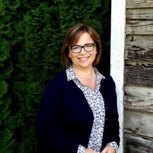 headshot of white woman in a dark sweater and paisley button down shirt amid a dark backdrop