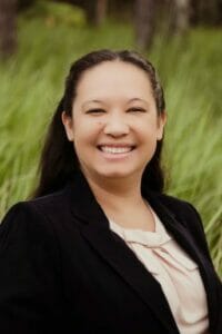 headshot of a hispanic woman with long black hair wearing a black blazer and light colored shirt with greenery in the background