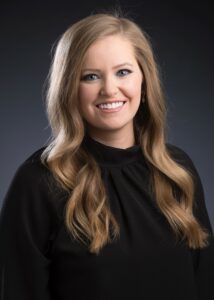 Headshot of white woman with long blonde hair wearing a black sweater amid a dark backdrop