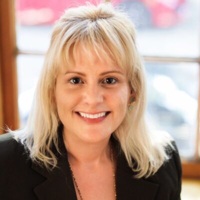 headshot of white woman with long blonde hair in a black blazer and purple shirt