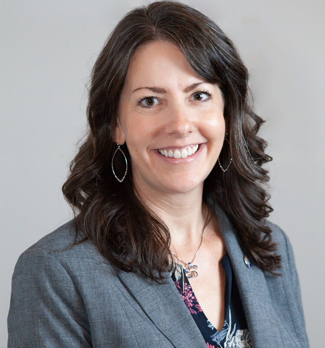 headshot of white woman with long brown hair smiling while wearing a grey blazer