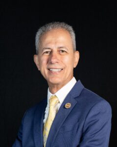hispanic man with grey hair in a pink tie, white collared shirt, and blue jacket amid a back backdrop