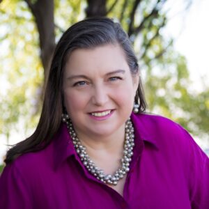 white woman with long brown hair with a natural background in a red shirt with a large necklace