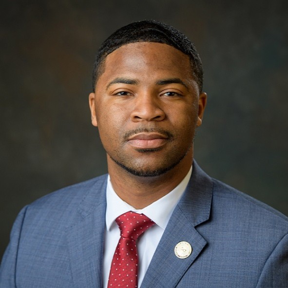 headshot of a black man in a light blue blazer, white collared shirt and red tie
