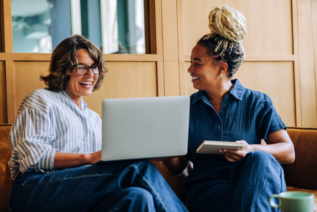 Two adult women enjoy a friendly discussion, using a laptop and a notebook. Their bright smiles convey positivity, teamwork, and productivity in a hotel lobby