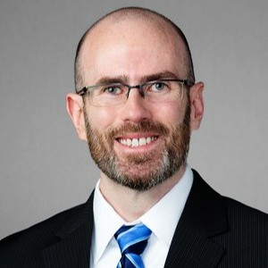 bearded white man wearing glasses and smiling wearing a black suit jacket, white collared shirt and blue tie
