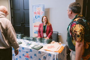Woman from MilMoneyCon talking to a man and a woman approaching the exhibit table at Symposium