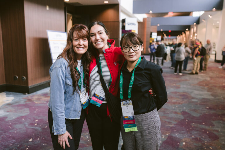 Three women, two wearing red Member ribbons, smiling at Symposium
