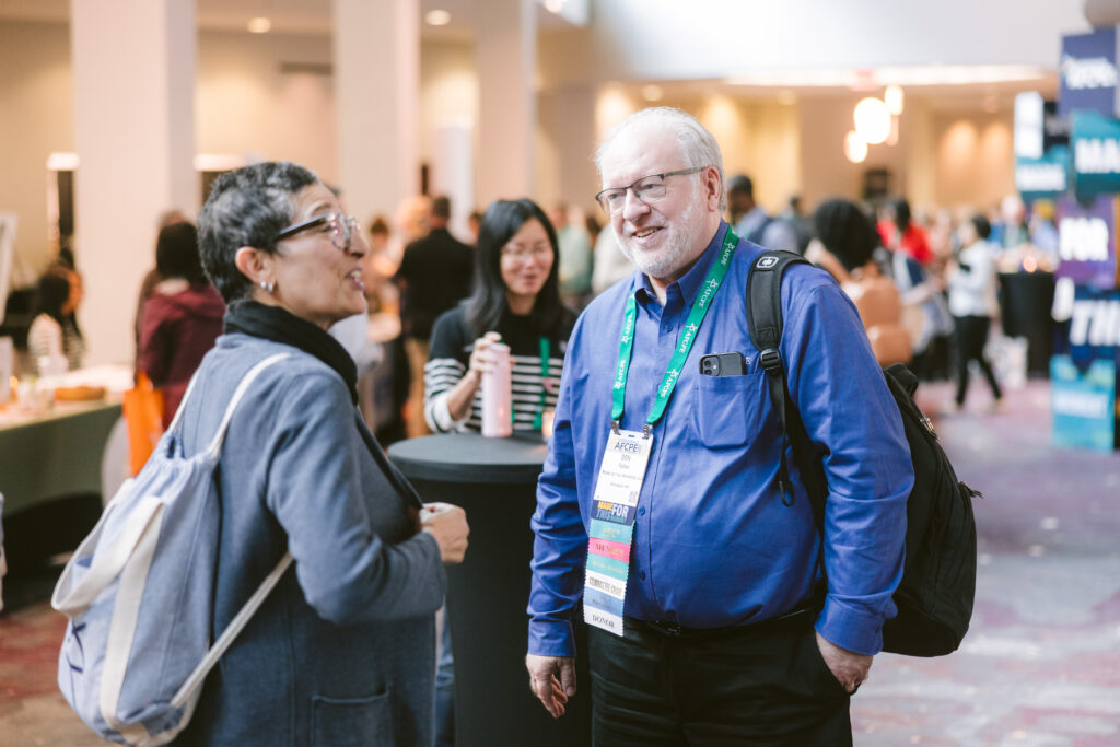 Older man and older woman having a conversation at Symposium. Older Man is in a blue shirt with a lanyard that has the red member ribbon.