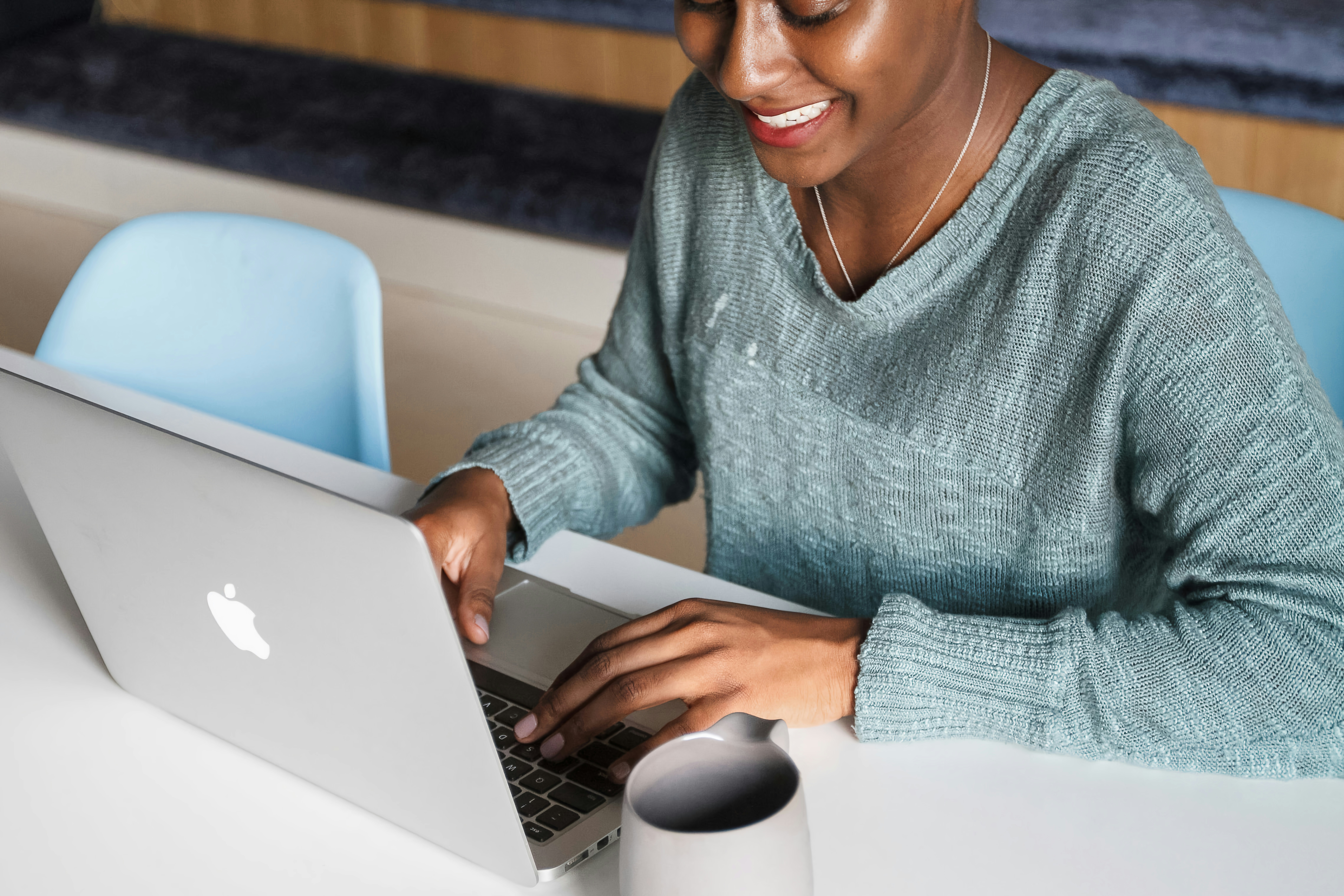 woman of color in a gray sweater typing on her laptop, coffee next to the laptop