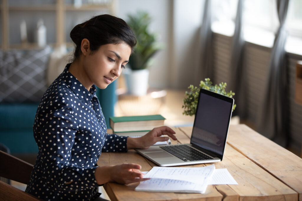 Millennial Indian girl sit at desk in living room study on laptop making notes, concentrated young woman work on computer write in notebook, take online course or training at home, education concept