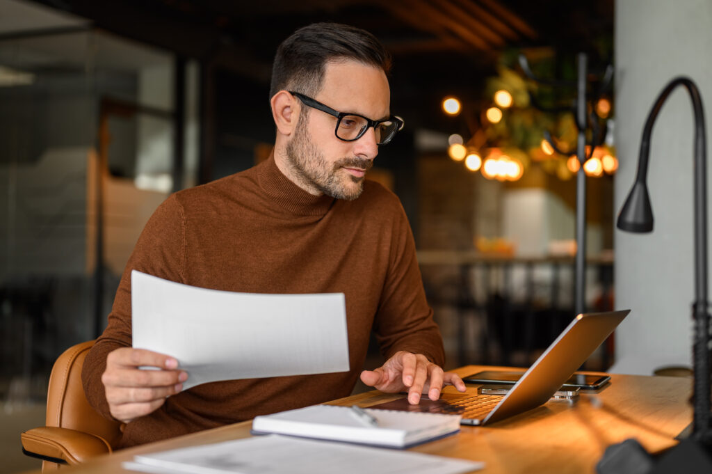 Focused business manager in eyeglasses auditing financial report and using laptop on desk in office