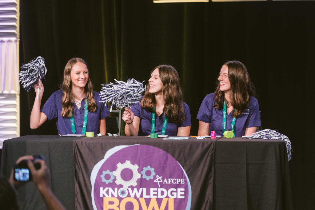 Three girl students waving pom poms behind their competition table at Knowledge Bowl at AFCPE Symposium