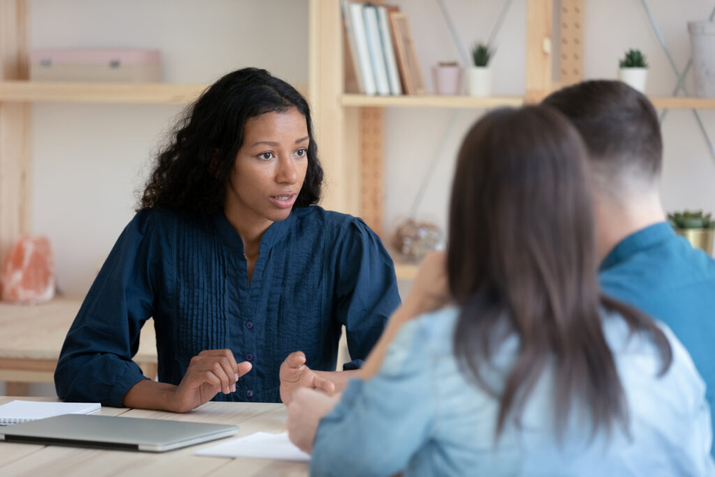 Focused African American female financial counselor talks with young couple at meeting, concentrated biracial woman consultant speak discuss ideas with clients spouses at office briefing