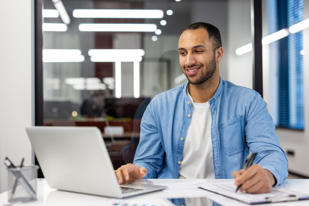 Smiling young male hispanic student sitting in modern office at desk with laptop and making notes in notebook, distance learning.
