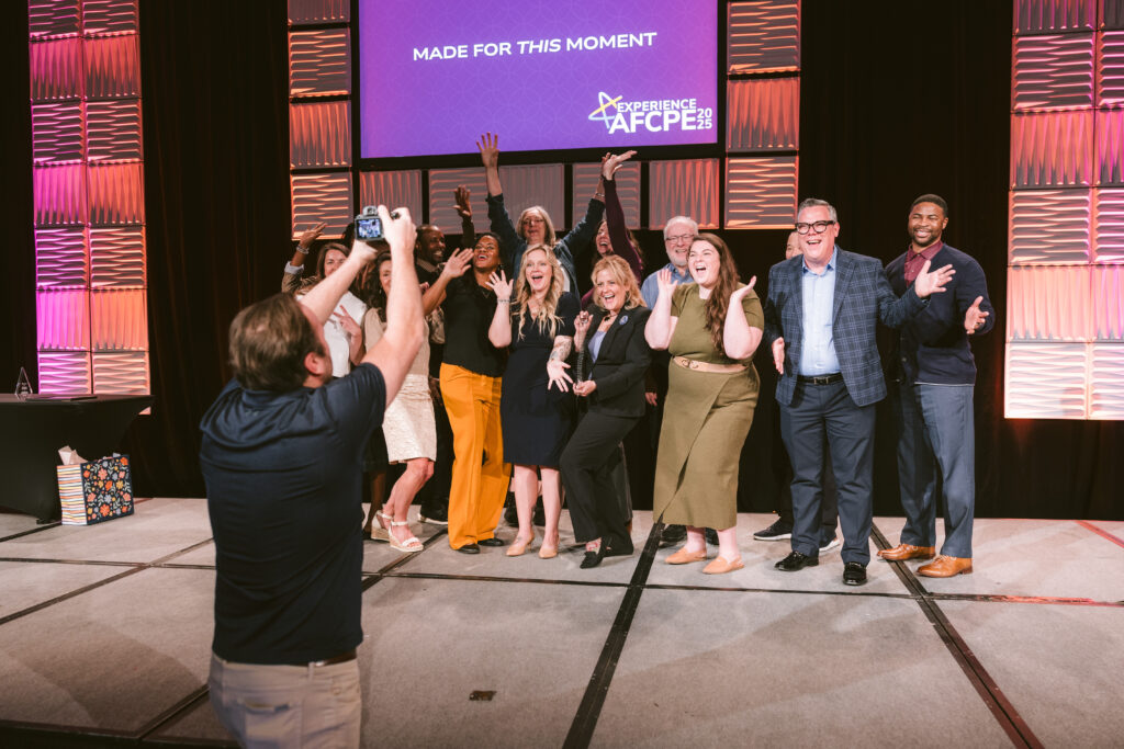 Photographer in a black shirt and khaki pants takes a picture of a diverse group of people on stage at Symposium