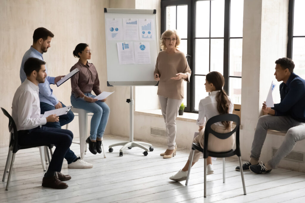 Confident middle-aged businesswoman lead meeting make flipchart presentation for diverse colleagues, motivated senior female coach present business project on whiteboard for coworkers in office