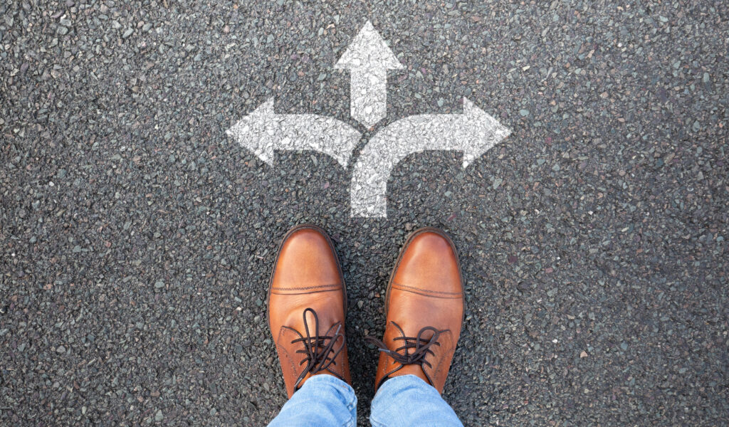 Two brown shoes of someone contemplating which direction they should go guided by three white arrows on the black pavement