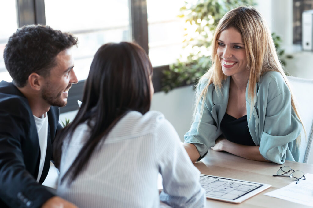 Shot of white woman with blonde hair wearing a light blue blazer discussing plans on electronic tablet while talking to her client couple.