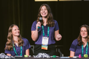 Three white college-aged women in matching purple polo shirts smiling, the one in the middle standing with a microphone