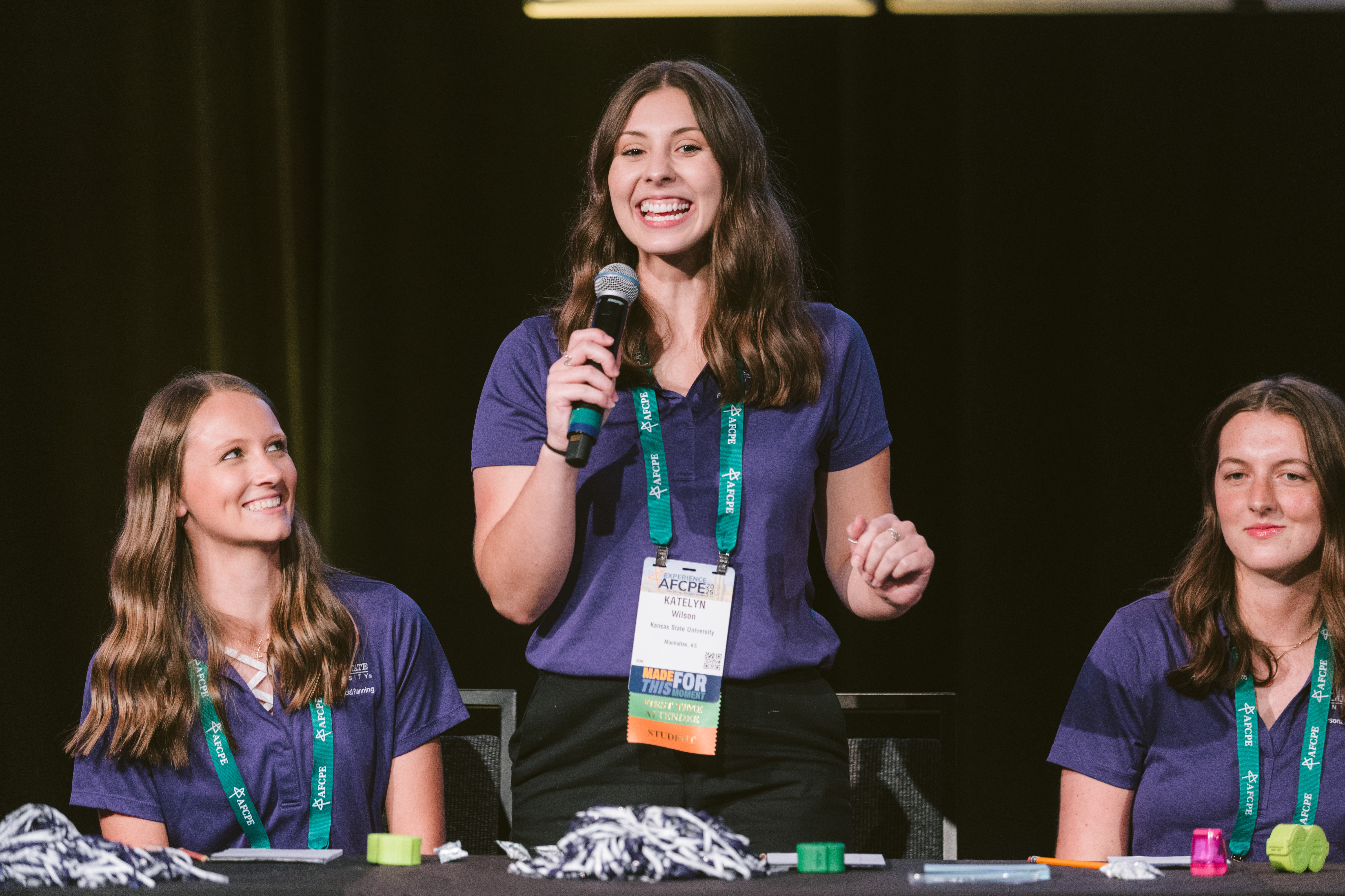 Three white college-aged women in matching purple polo shirts smiling, the one in the middle standing with a microphone