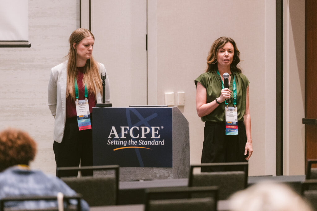 a white woman holding a microphone while another white woman watches during a presentation