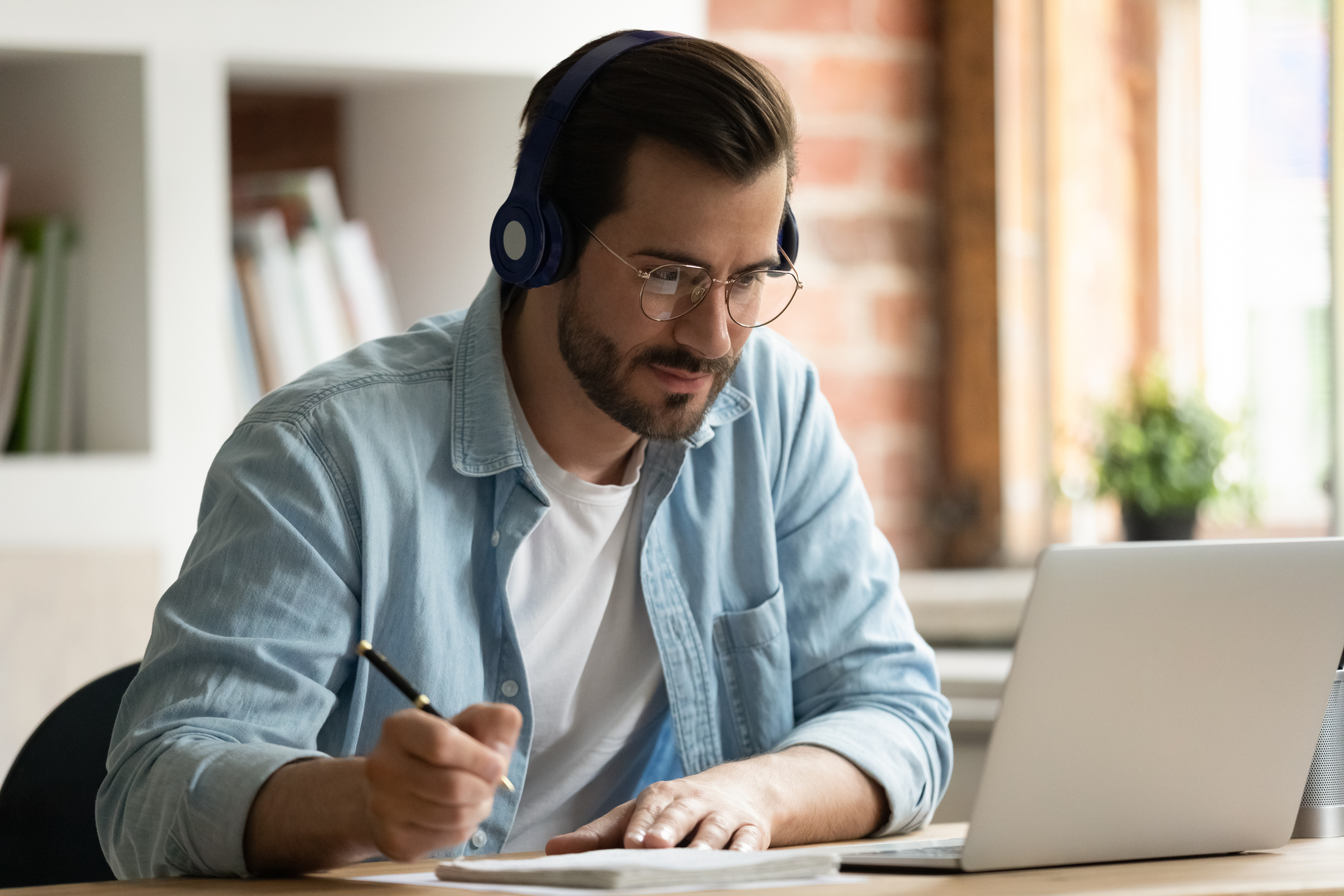 Happy focused young man in eyeglasses wearing wireless headphones, watching educational lecture on computer, studying on online courses distantly, improving professional knowledge, writing notes.