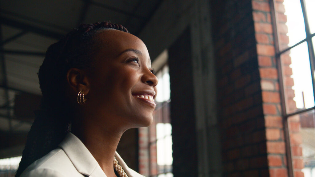 A portrait of a confident, smiling woman standing near industrial-style windows in a modern architecture setting. The natural light accentuates her joyful expression, capturing a sense of positivity and professional success.