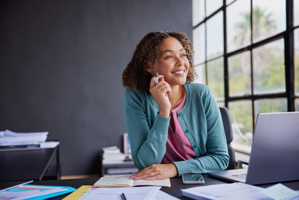 Smiling beautiful african american woman sitting at her desk while looking thoughtful and inspired. Professional business woman taking a moment to reflect. Cheerful businesswoman holding a pen while thinking about the future, enjoying a quiet moment in modern office.