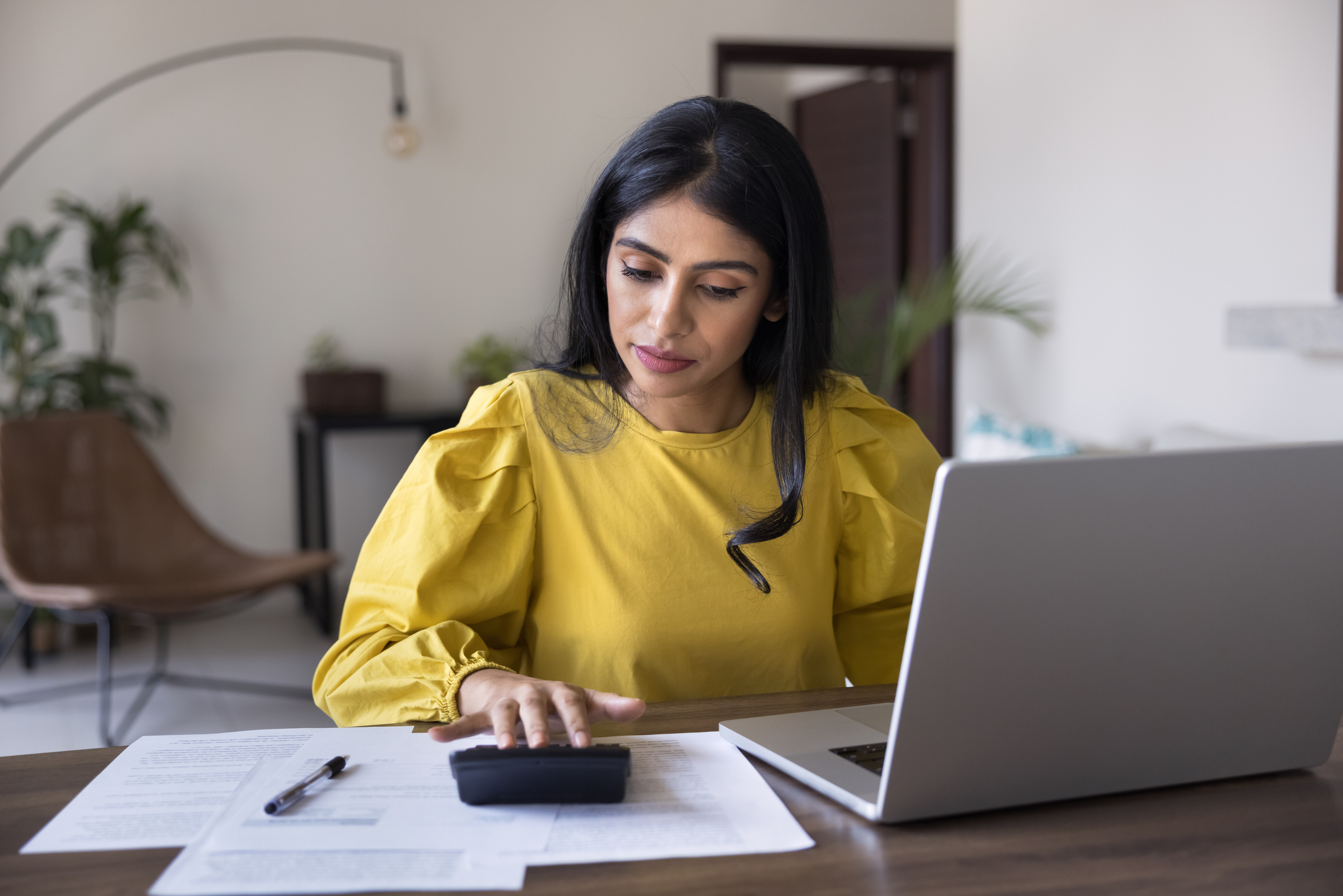 Focused Indian woman sitting at desk with laptop, using calculator, calculates expenses, managing financial records, organizing monthly incomes and expenses, budgeting, checking documents for payments