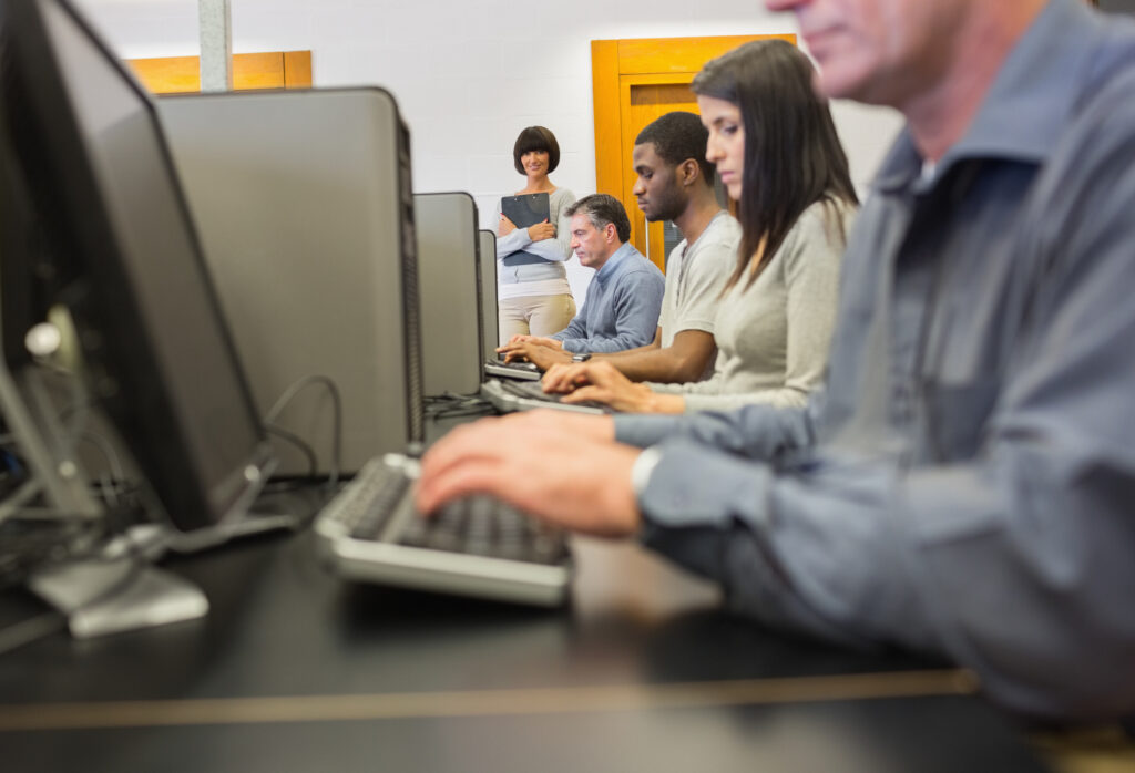diverse group of adult learners taking an online test overseen by a proctor in the distance