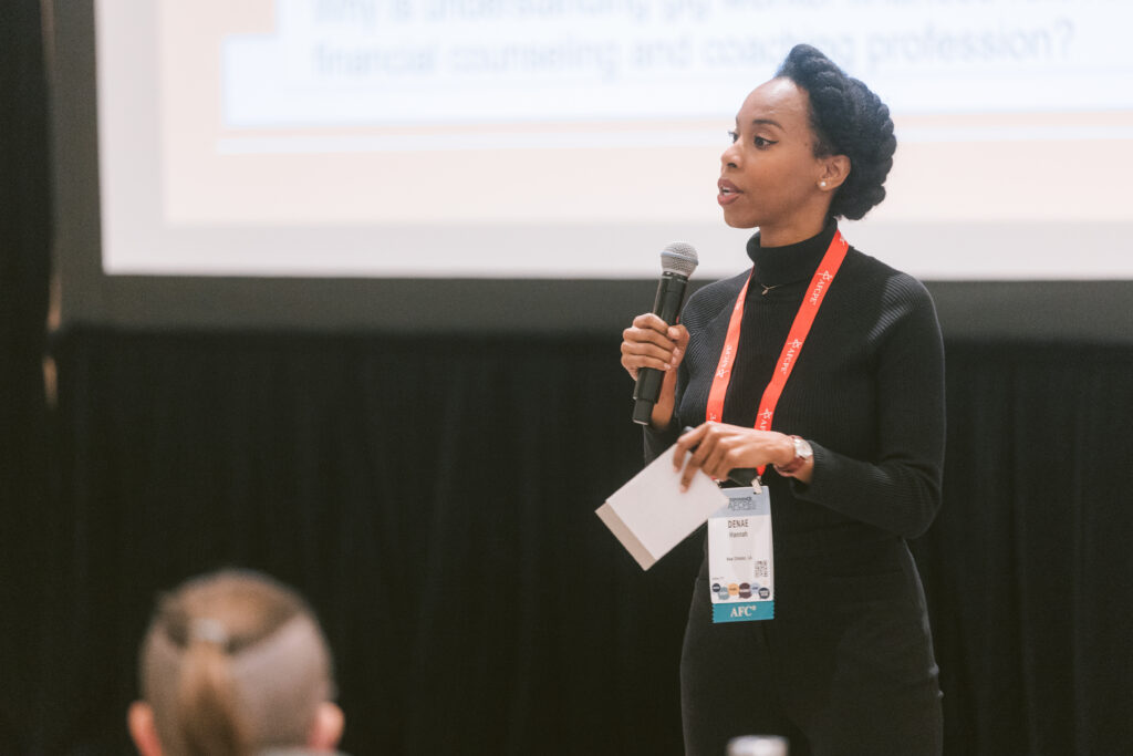 black woman wearing all black holds a microphone while answering questions during a presentation
