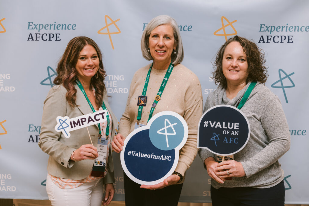 three white woman of varied ages smiling and holding AFC and AFCPE signs at Symposium