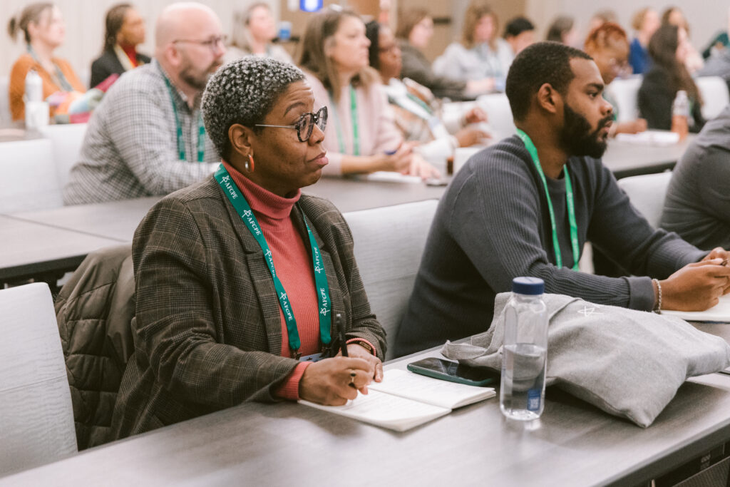 focused black woman taking notes during a presentation. To her left, a black man is listening to the presentation.