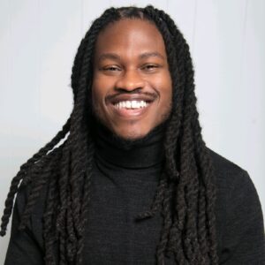 black man with long braided brown hair and a mustache smiling and wearing a dark turtleneck sweater