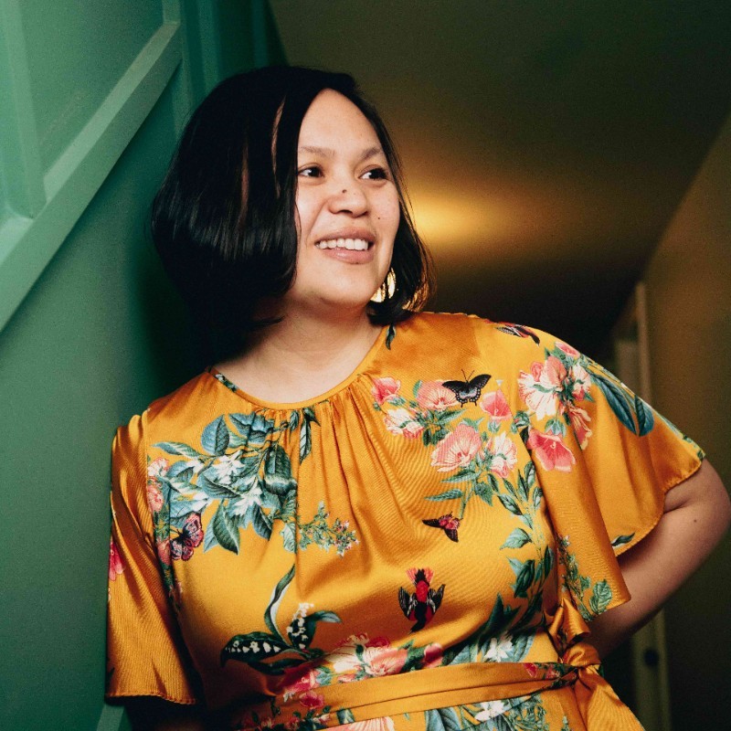 asian woman with shoulder length black hair wearing a patterned orange shirt smiling while looking to the side