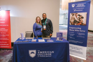 A man and woman standing at an American College exhibitor table at Symposium