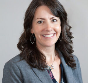 headshot of white woman with long brown hair smiling while wearing a grey blazer