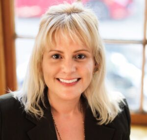 headshot of white woman with long blonde hair in a black blazer and purple shirt