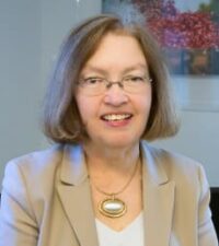 white woman with shoulder length grey hair in a beige blazer and glasses with a white shirt