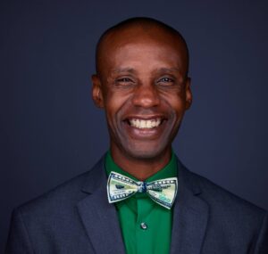 Dark background, man of color, light colored bow tie, green shirt, dark blazer