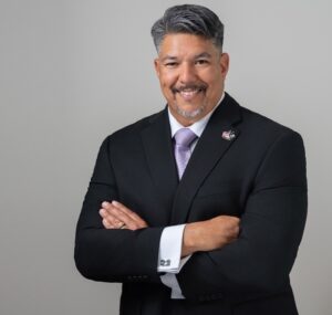 hispanic man smiling with grey hair and a grey goatee wearing a dark suit, white collared tie, and salmon tie